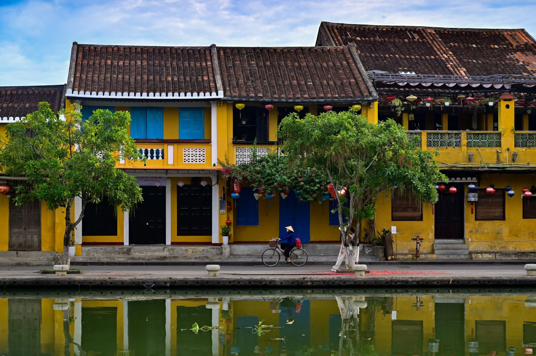 A cyclist passes yellow historic houses beside the water in Hoi An